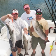 Three smiling men standing on a boat deck on a sunny day. The man on the left and the man on the right are holding up large striped bass they have caught, while the man in the center stands between them. Several fishing rods in holders and Yamaha boat motors are visible in the background.