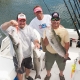 Three smiling men standing on a boat deck on a sunny day. The man on the left and the man on the right are holding up large striped bass they have caught, while the man in the center stands between them. Several fishing rods in holders and Yamaha boat motors are visible in the background.