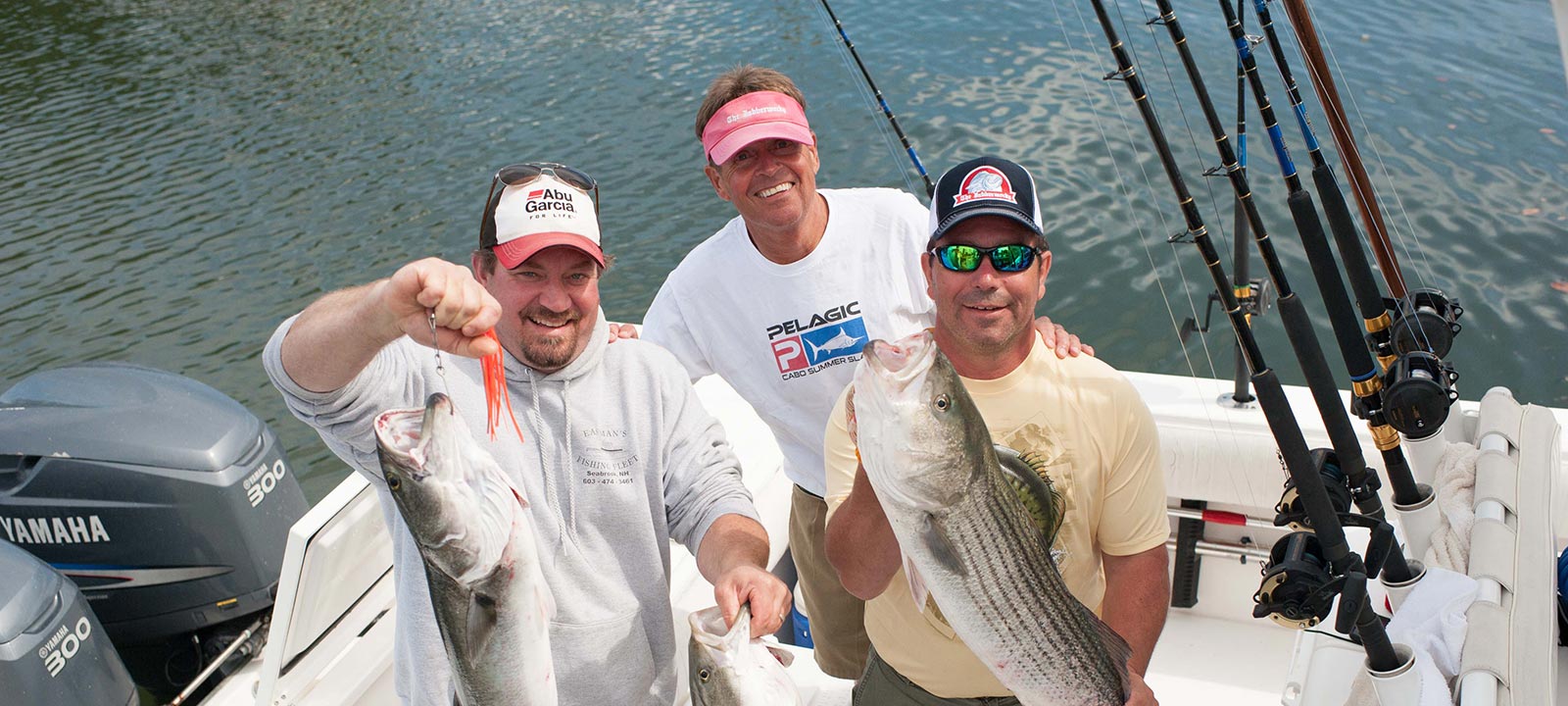 Three smiling men standing on a boat deck on a sunny day. The man on the left and the man on the right are holding up large striped bass they have caught, while the man in the center stands between them. Several fishing rods in holders and Yamaha boat motors are visible in the background.